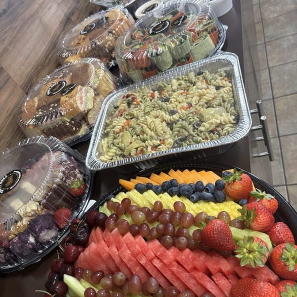A deli catering spread with pasta salad, fruit platter, sandwiches, and wraps in clear containers.