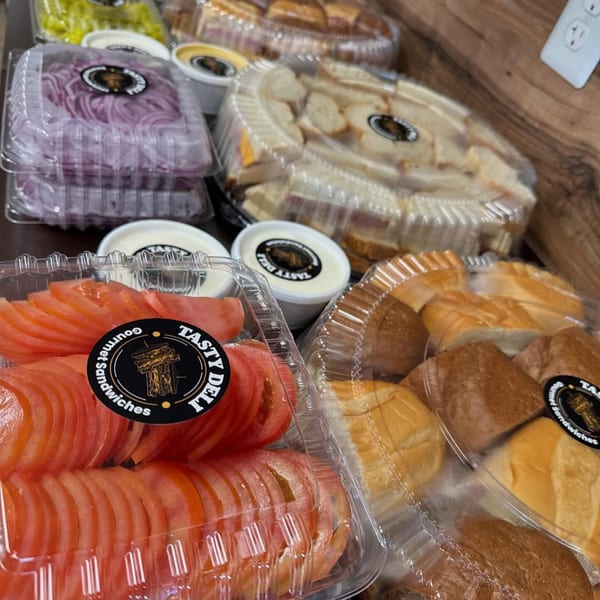 Tasty Deli catering trays with sliced tomatoes, red onions, bread rolls, and sandwiches.