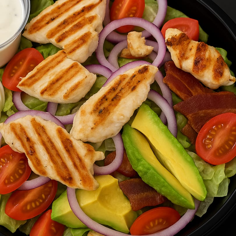 Grilled chicken salad with lettuce, tomatoes, red onion, avocado, bacon, and a side of dressing.